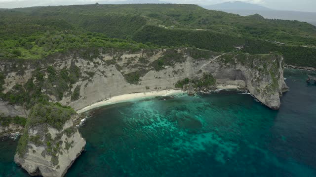 空中倾斜到热带海滩海湾坐落在陡峭的悬崖边上，热带绿松石的水，珊瑚，岩石，和拍打的海浪-努沙佩尼达和龙目岛，巴厘岛视频素材