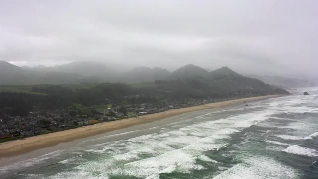 空中倾斜向下拍摄的海浪溅在海滩上的城市与天空，无人机飞向靠近山脉的海岸线-坎农海滩，俄勒冈州视频素材