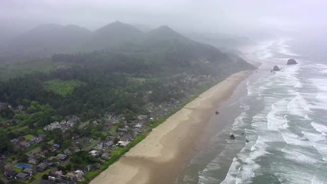 航拍海浪溅在海滩上的城市和山脉，无人机向前飞行的海岸线对天空-坎农海滩，俄勒冈州视频素材