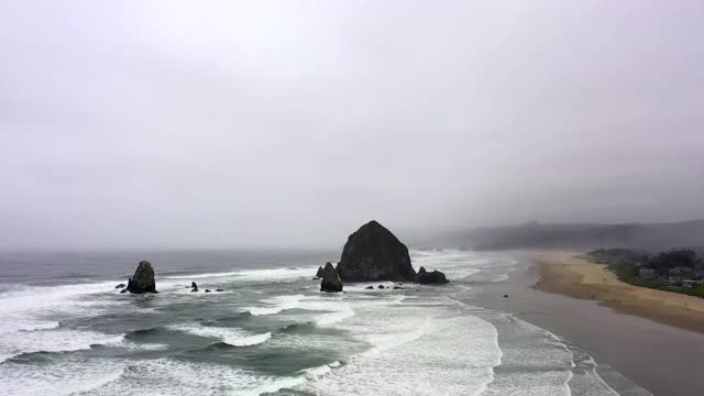 航拍海浪溅在海滩上对多云的天空，无人机上升在城市附近的海岸线-坎农海滩，俄勒冈州视频素材