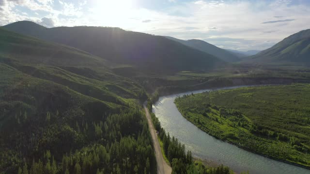 空中平移拍摄的道路在阳光明媚的一天河流对山脉，无人机飞过绿色的森林对天空-大北方平原，MT视频素材