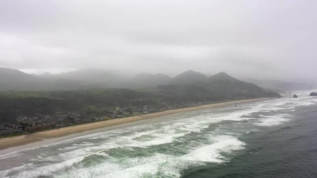 航拍海浪溅在海滩上的城市对天空，无人机在靠近山脉的海岸线上飞行-坎农海滩，俄勒冈州视频素材