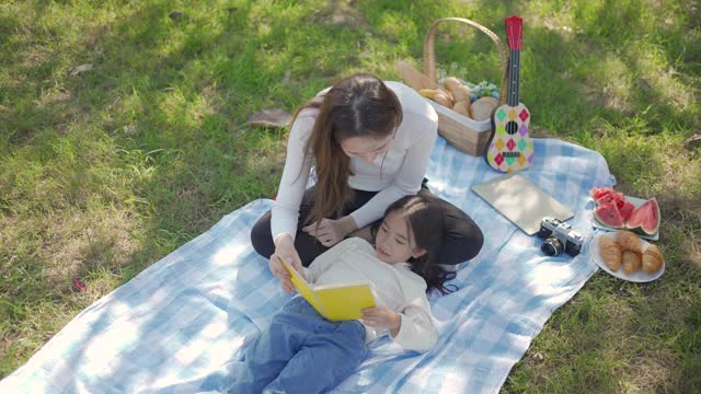 美丽年轻的亚洲母亲和女儿野餐和阅读黄色书在绿色公园。父母在暑假教孩子。人们的生活方式和幸福的家庭生活。视频素材