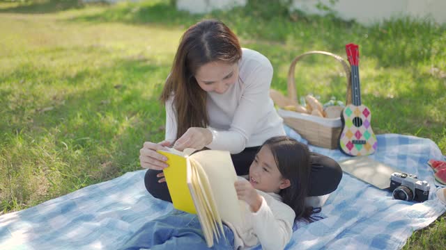 美丽年轻的亚洲母亲和女儿野餐和阅读黄色书在绿色公园。父母在暑假教孩子。人们的生活方式和幸福的家庭生活。视频素材