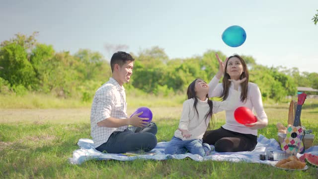 幸福的家庭亚洲父亲，母亲和小女孩玩气球，并享受了我们一起在野餐布在绿色的花园。一家人在大自然中享受阳光明媚的秋日。视频素材