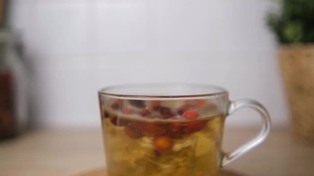 用玫瑰果和莱姆花泡茶。水果茶。在杯子里冲泡。在厨房的背景——一个花盆和一朵花。散景。模糊的背景。软木板。4 k。视频素材