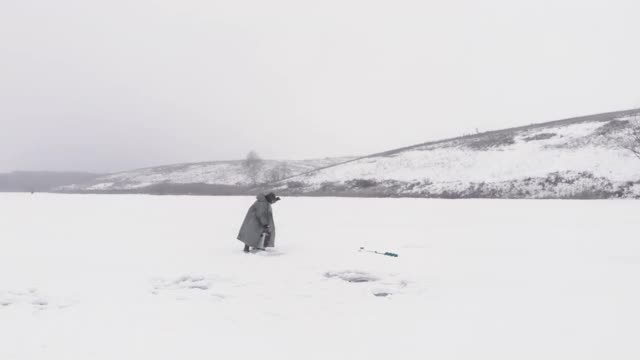 人类独自在冰冻的河上钓鱼。冬季捕鱼的概念。渔夫用钓竿在结冰的湖或池塘的冰洞里捕鱼。空中无人机拍摄的视频素材