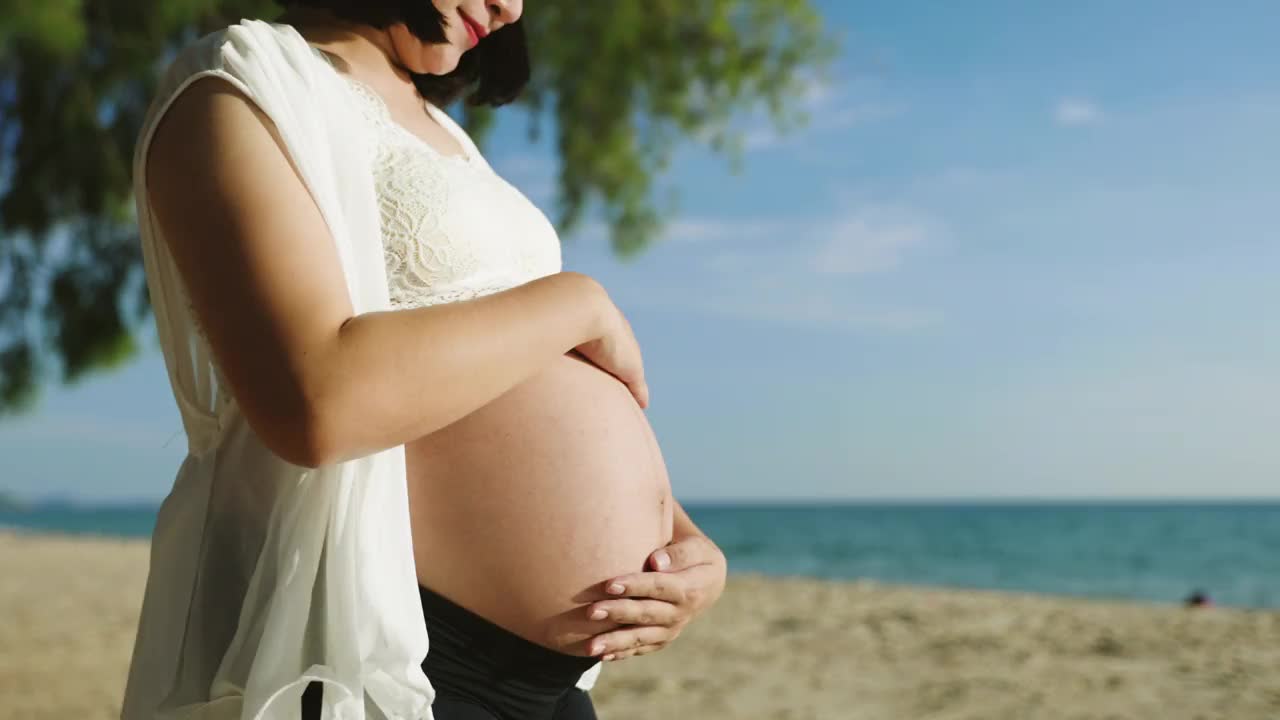 近距离观察年轻的亚洲孕妇触摸她的肚子期待宝宝健康放松在海滩上视频素材