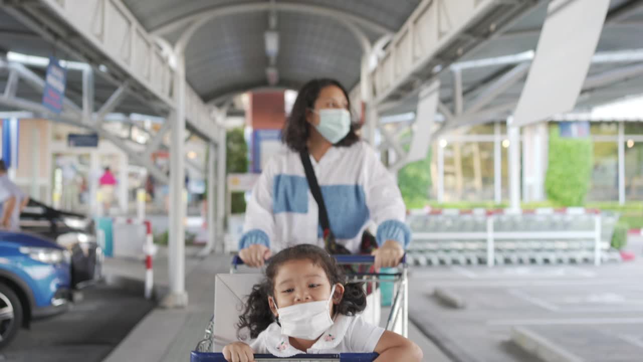 亚洲母亲与可爱的孩子女孩戴着防护口罩推购物车在停车场视频素材