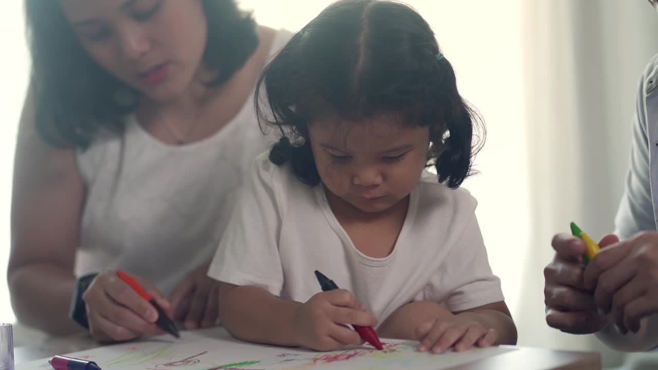 亚洲爸爸和妈妈一起在客厅教小女儿用彩色铅笔画和在纸书上画画视频素材