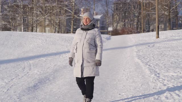 在阳光明媚的日子里，一名孕妇穿着冬季保暖夹克，带着兜帽漫步在白雪覆盖的公园里视频素材