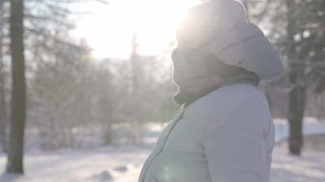 在阳光明媚的日子里，一名孕妇穿着冬季保暖夹克，带着兜帽漫步在白雪覆盖的公园里视频素材