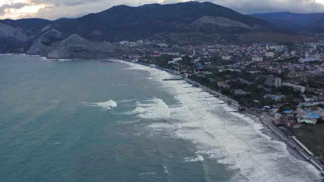 海滨的现代城市以群山和黄昏的天空为背景。城市海滩上的海浪。从无人机上方俯瞰度假城市和山区景观。4 k鸟瞰图视频素材