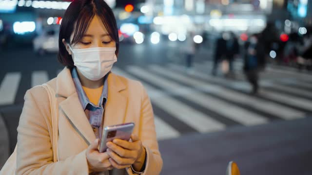 年轻女子用保护性面罩遮住脸部，晚上在城市街道上使用电话视频素材