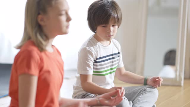 肖像集中的白人男孩坐在莲花的姿势看模糊的女孩在前面。在家里和朋友或姐妹一起集中训练深色头发的孩子。运动与宁静的概念。视频素材