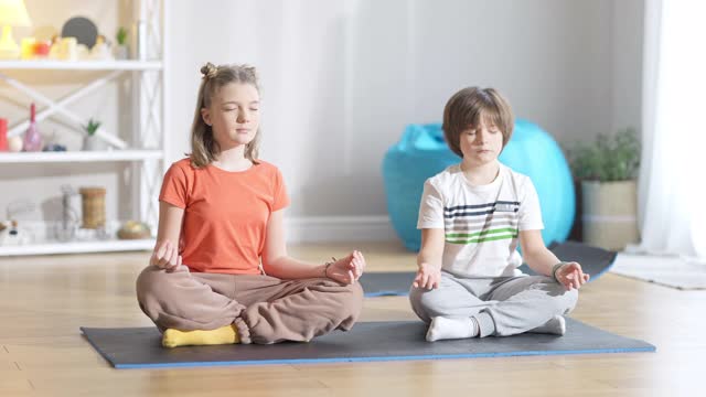 集中的白人女孩坐在运动垫子上闭上眼睛，男孩重复瑜伽训练后的兄弟姐妹。小瑜伽行者在家锻炼的广角镜头。运动和幸福的概念。视频素材