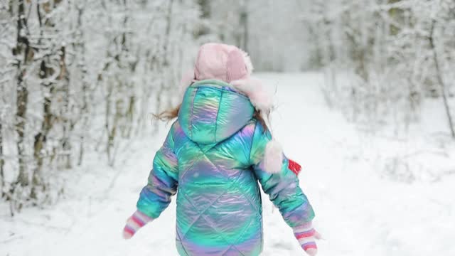 后视图快乐可爱的孩子女孩在一个缤纷的衣服和帽子在一个下雪的冬天公园跑步。冰雪森林里的冬姑娘。漫步冬日森林100fps视频素材