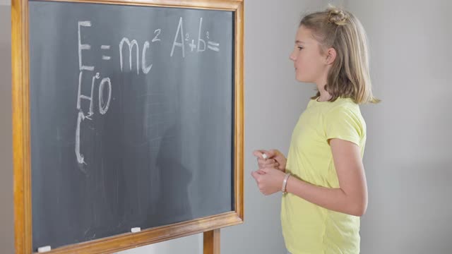 漂亮聪明的白人女学生在黑板上摆姿势写着物理公式。可爱自信的天才女孩看着相机微笑的肖像。智慧教育理念。视频素材