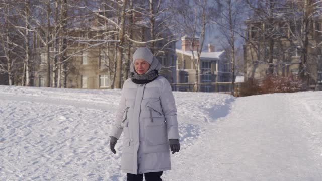 在阳光明媚的日子里，一名孕妇穿着冬季保暖夹克，带着兜帽漫步在白雪覆盖的公园里视频素材