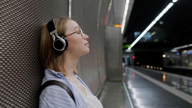 一位年轻的现代女孩坐在地铁的长椅上等车，耳机里戴着耳机听着音乐视频素材