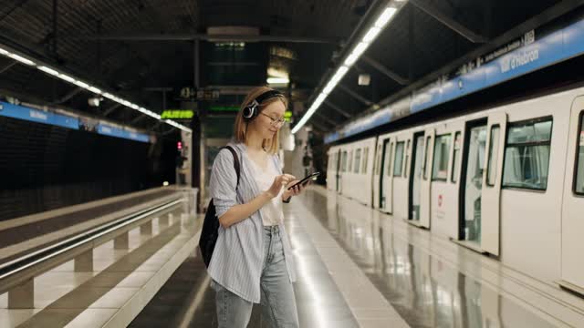 女孩在地铁站等火车，戴着耳机听音乐。在地铁里使用智能手机。地下。学生乘坐地铁视频下载