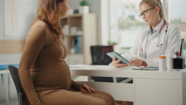 女医生在诊所咨询时向年轻的准妈妈展示平板电脑和超声妊娠扫描。坐在医院办公室桌子后面的家庭医生。视频素材