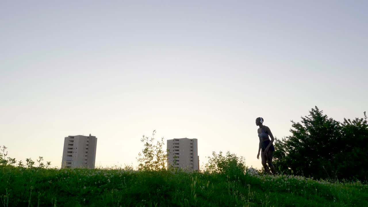 日落时分，一个戴着耳机的女人在草地上慢跑的慢镜头视频素材