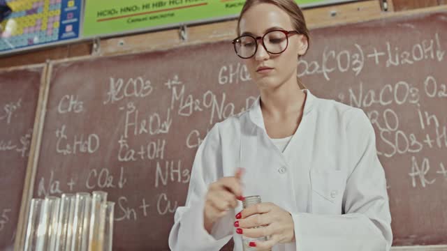 年轻女子戴着眼镜，身穿白袍，打开瓶子，将液体加入蓝色试剂中，并在黑板上用公式进行化学实验视频素材