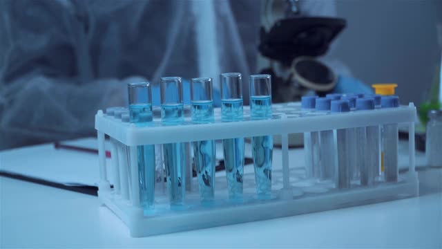 生化实验室研究，穿着防护服的化学家在实验室用仪器、显微镜和含有化学液体的科学实验玻璃器皿分析蓝色样品。视频素材