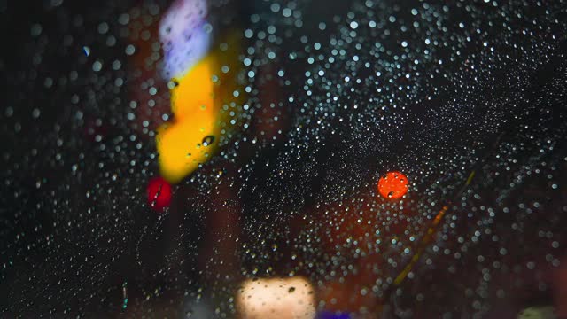 雨点在夜晚城市明亮的灯光背景下从汽车玻璃上流下。雨夜。汽车通过十字路口。鲜艳的交通灯。视频素材