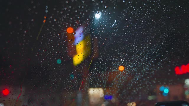 雨点在夜晚城市明亮的灯光背景下从汽车玻璃上流下。雨夜。汽车通过十字路口。鲜艳的交通灯。视频素材