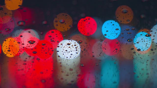 雨点在夜晚城市明亮的灯光背景下从汽车玻璃上流下。雨夜。汽车通过十字路口。鲜艳的交通灯。视频素材