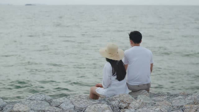 男人和女人夫妇坐在海边的石头上享受假期，夏天愉快地说着，笑着，在晚上看海滩上的景色。视频素材