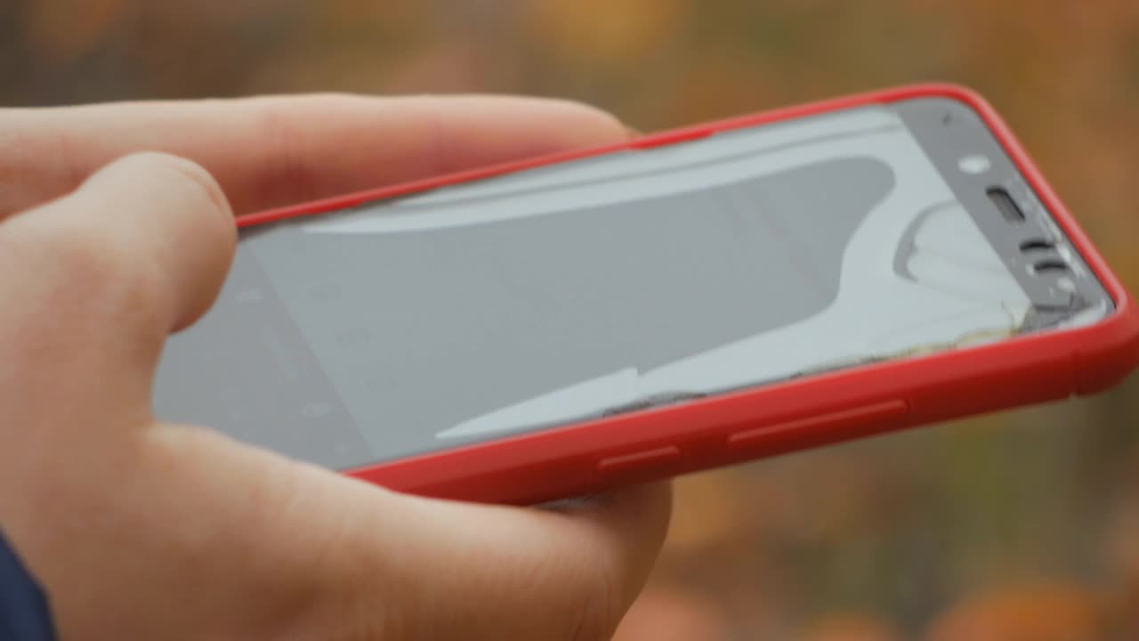 红色手机屏幕破碎。近距离的女人的手触摸红色手机与模糊的黄绿色背景，发送短信。视频下载