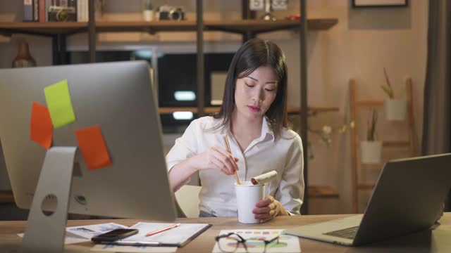 亚洲聪明的自由职业商业女性晚上在家里的客厅里用笔记本电脑工作的时候吃着方便面。股票视频素材