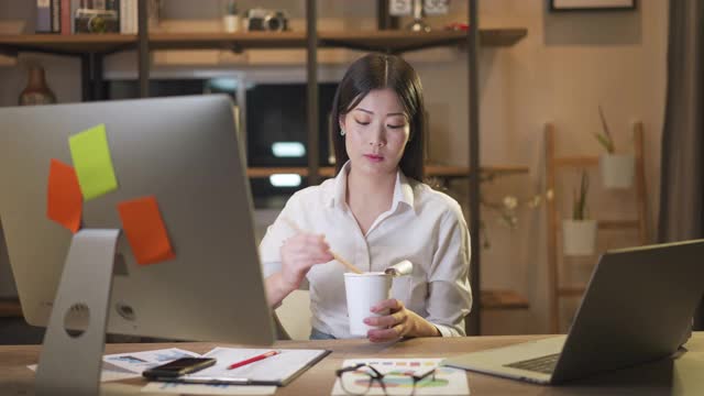 亚洲聪明的自由职业商业女性晚上在家里的客厅里用笔记本电脑工作的时候吃着方便面。股票视频素材