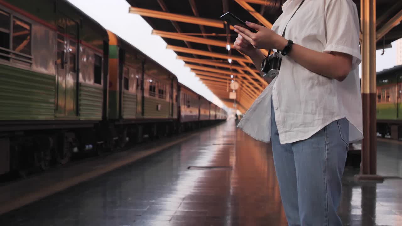 旅行家背包亚洲女人旅行在曼谷，泰国。快乐的年轻女性坐在长凳上用智能手机聊天，阅读和发短信在火车站。视频素材