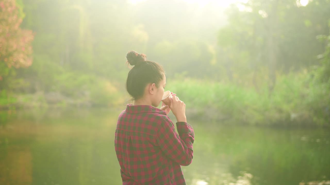 一个美丽的年轻旅行者女人是享受与自然喝咖啡在早晨湖，放松，露营假日和旅游概念。视频素材