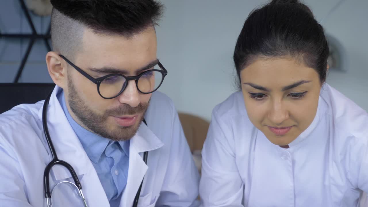 Two doctors, a Man and a Woman, Looking at a Tablet screen or Working at a Computer or Laptop, Are in a Bright Hospital Office .两个医生的特写，一个男人和一个女人，看一个平板电脑屏幕或工作在电脑或笔记本电脑，在一个明亮的医院办公室视频素材