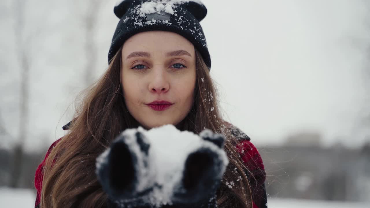 在冬天的公园里，穿着暖和的冬衣和帽子的女人在吹雪。飞的雪花。阳光明媚的一天。快乐的美丽年轻女孩有乐趣在霜冻的森林视频素材