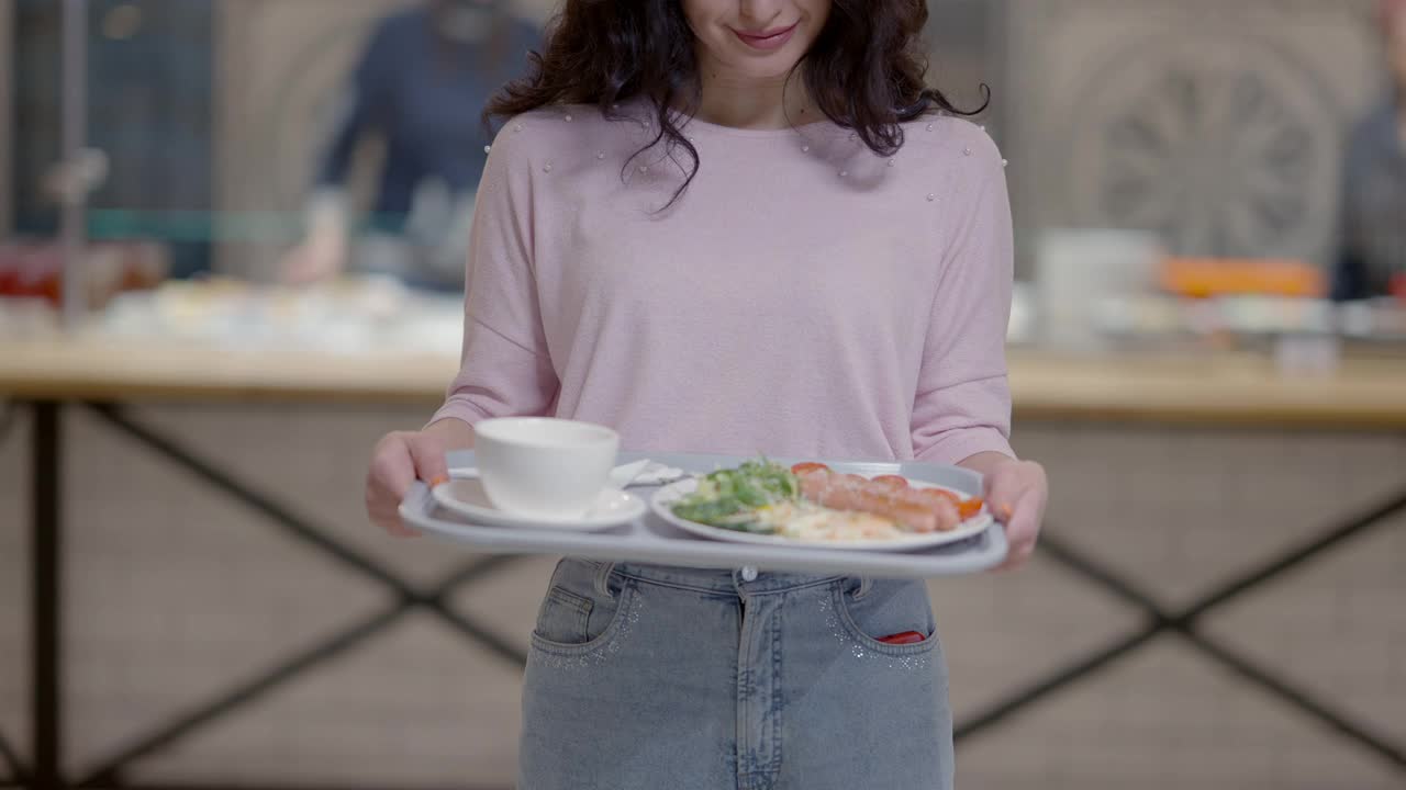 一个面目人非的苗条年轻女子，手里拿着盛着食物和茶的托盘，面带微笑。积极的白人女士摆姿势在自助餐厅与早餐或午餐。餐饮和生活方式。视频素材