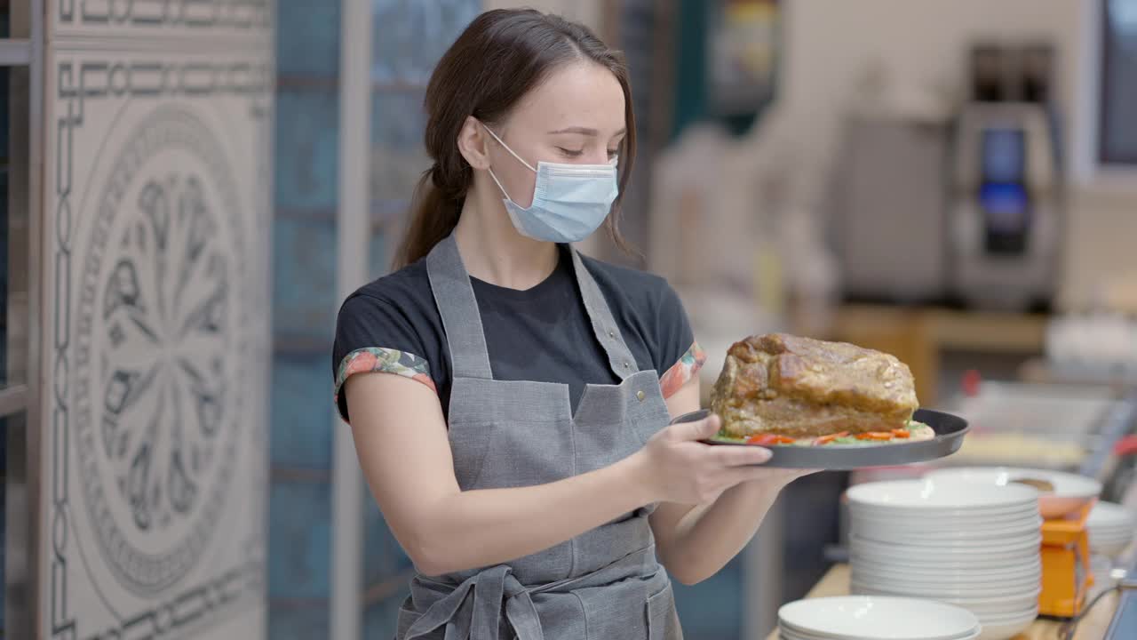 年轻漂亮的白种人厨师或女服务员戴着口罩在餐厅用餐。积极苗条的女人拿着熟肉微笑。冠状病毒新常态的生活方式和烹饪。视频素材