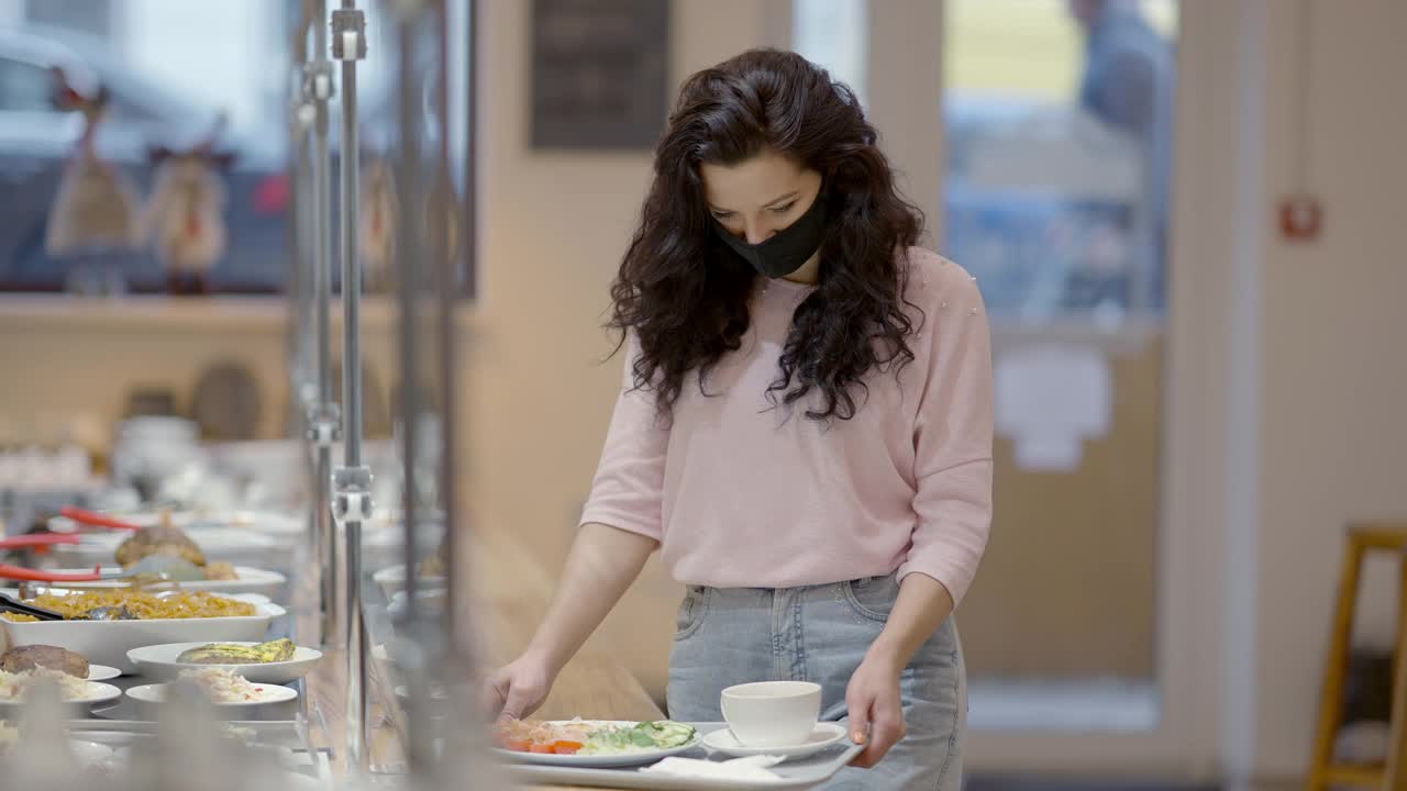 戴着新冠肺炎口罩的年轻白人女性在自助餐厅选择食物，然后慢镜头离开。一位自信的女士在餐厅吃早午餐的照片。视频素材