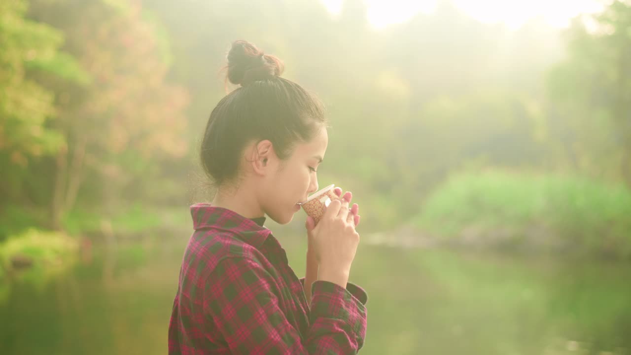 一个美丽的年轻旅行者女人是享受与自然喝咖啡在早晨湖，放松，露营假日和旅游概念。视频素材