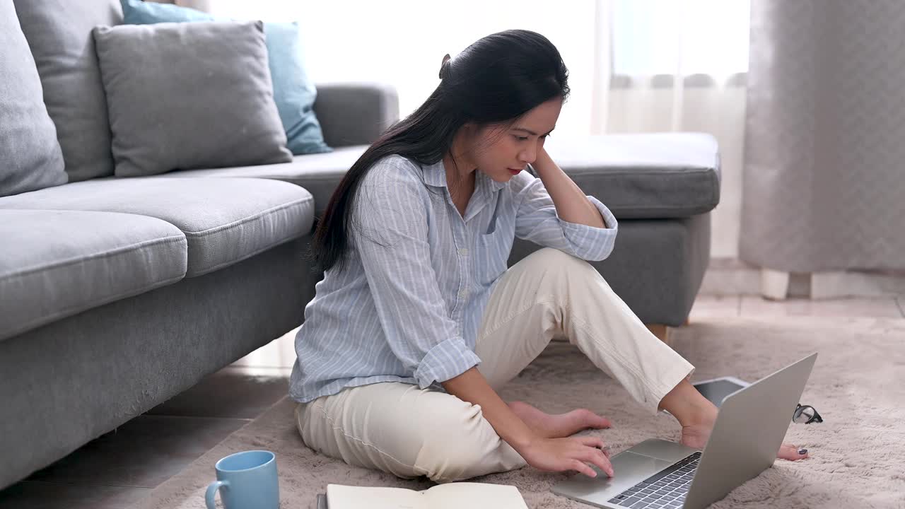 一位年轻的亚洲女商人正在笔记本电脑上寻找信息，她在笔记本上做笔记，她坐在家里客厅的地毯上。商业女性和工作理念。视频素材