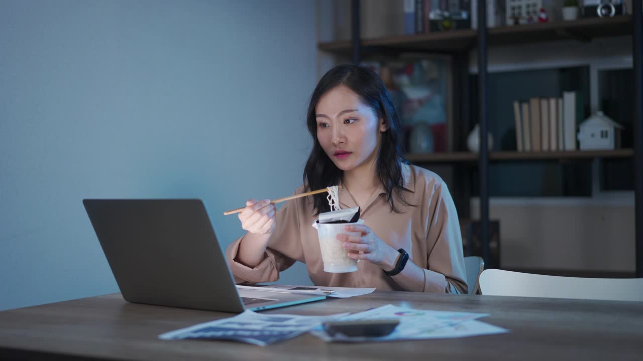 亚洲聪明的自由职业商业女性晚上在家里的客厅里用笔记本电脑工作的时候吃着方便面。视频素材