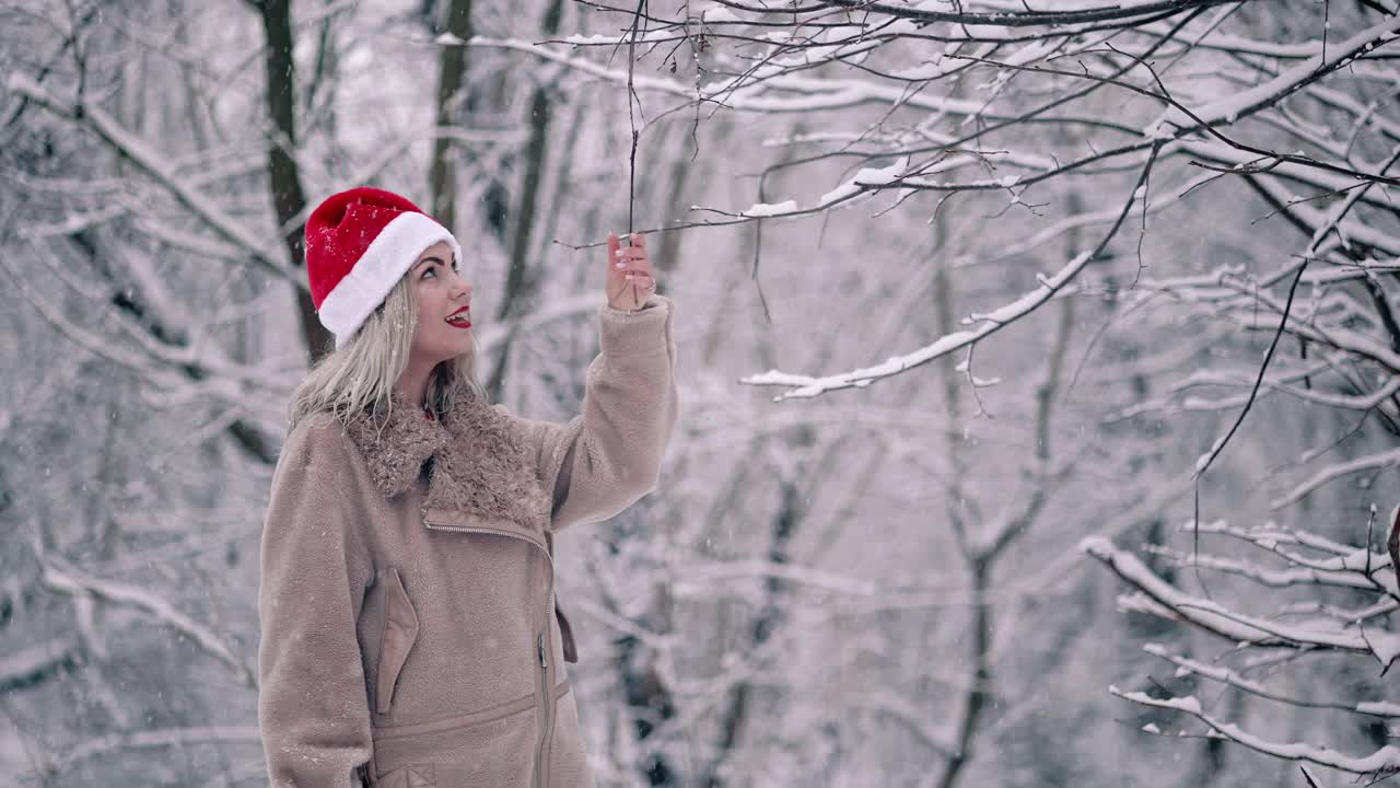 快乐可爱的女人戴着圣诞老人的帽子拉上树枝，雪花飞在她的身上，她快乐地欢庆冬天和寒冷的天气。缓慢的运动。视频素材