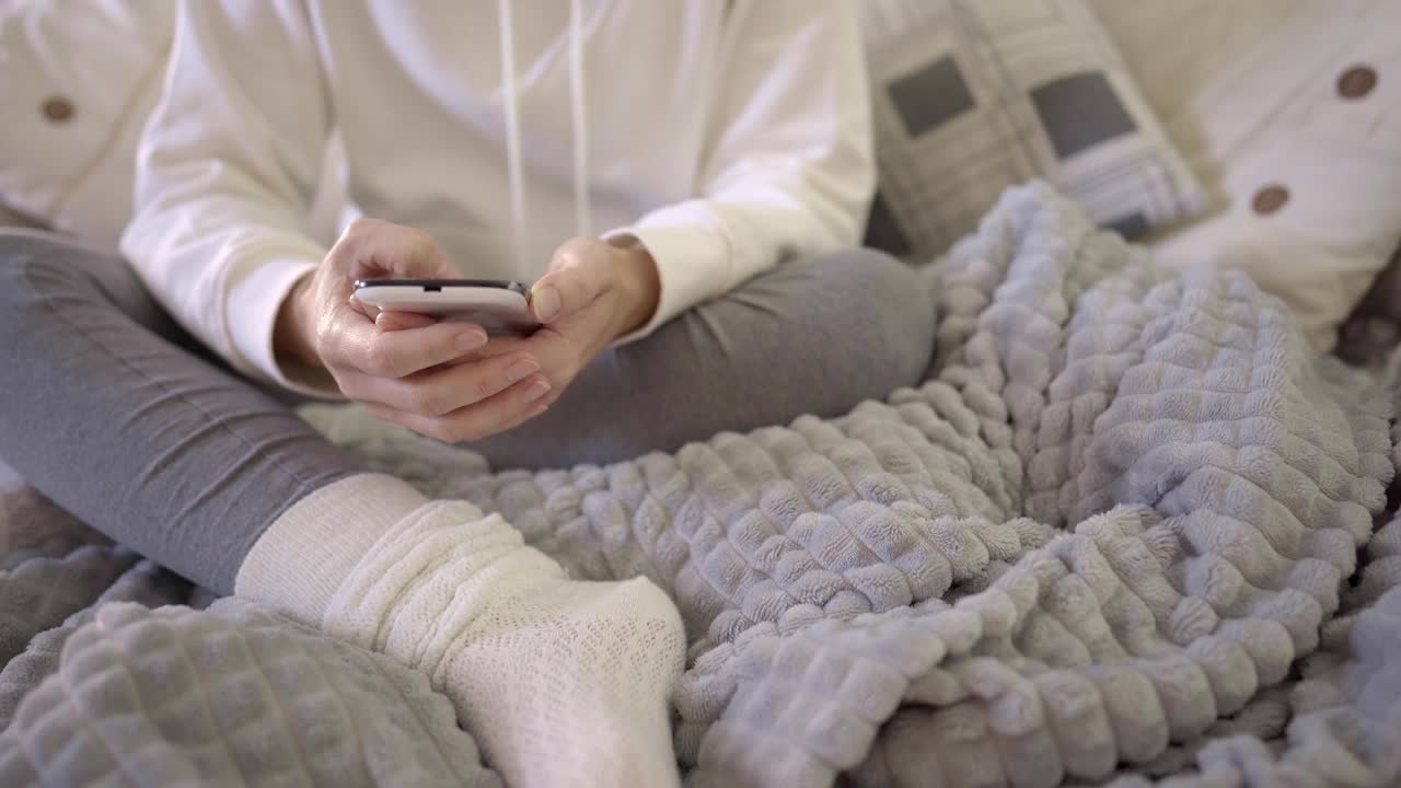 一个面面相觑的年轻女子被迫呆在家里，花时间坐在沙发上，用她的智能手机上网、网上购物，在社交网络上分享。女性千禧在沙发上与细胞视频素材