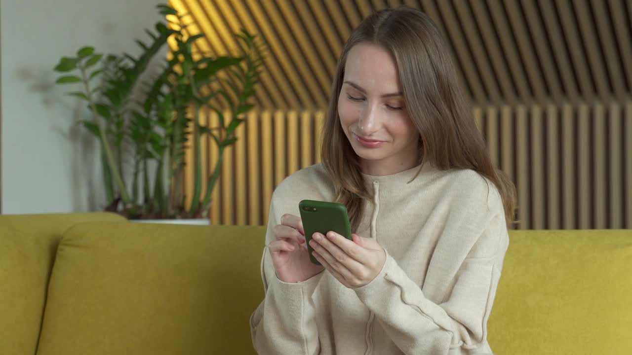 一名女子坐在客厅的黄色沙发上使用智能手机。一名妇女在家里用智能手机上网视频素材