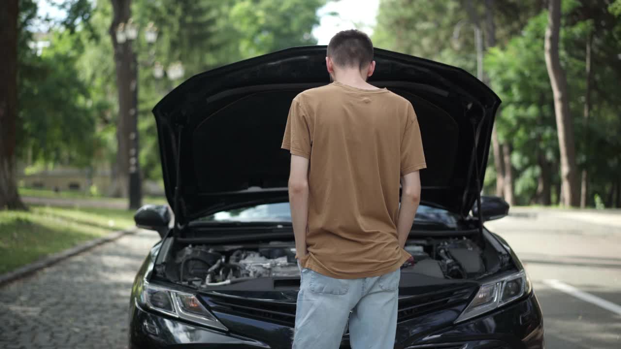 在夏天的道路上，年轻的白人男子紧握着头看着打开的汽车引擎盖。一个在室外发生交通事故的人。破碎和维修理念。视频素材